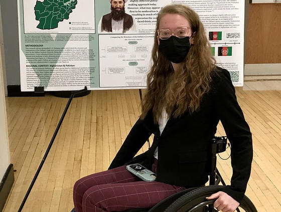 A young woman in eye glasses and a black mask sits in a wheelchair in front of a poster she created about the Taliban.