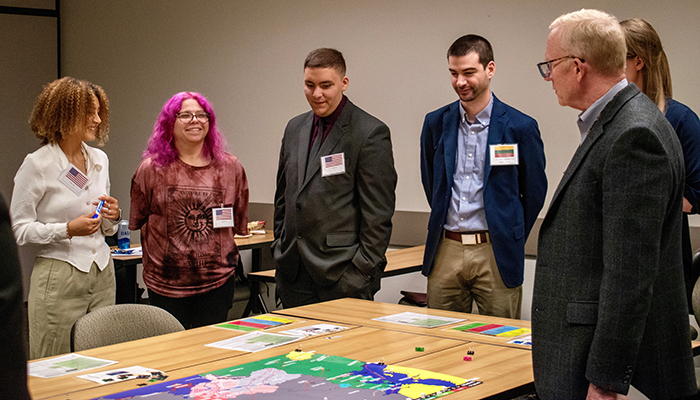 Five students stand around a table with a colorful board game on it as a man in a suit and glasses watches.