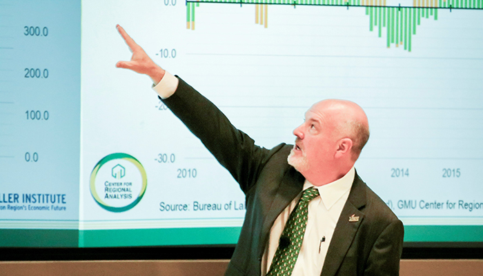 A man in a suit and green tie points to numbers on a screen behind him.