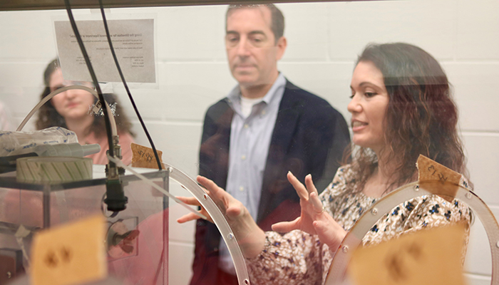A woman gestures with her hands behind a Plexiglas box as others look on.