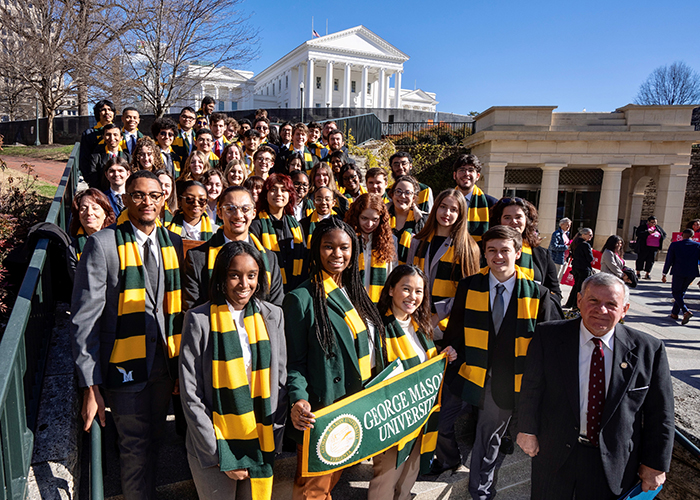 A group of young people wearing green and gold winter scarfs stand in front of a white government building.