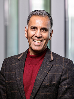 A man in a maroon mock turtleneck sweater and a brown jacket smiles at the camera.