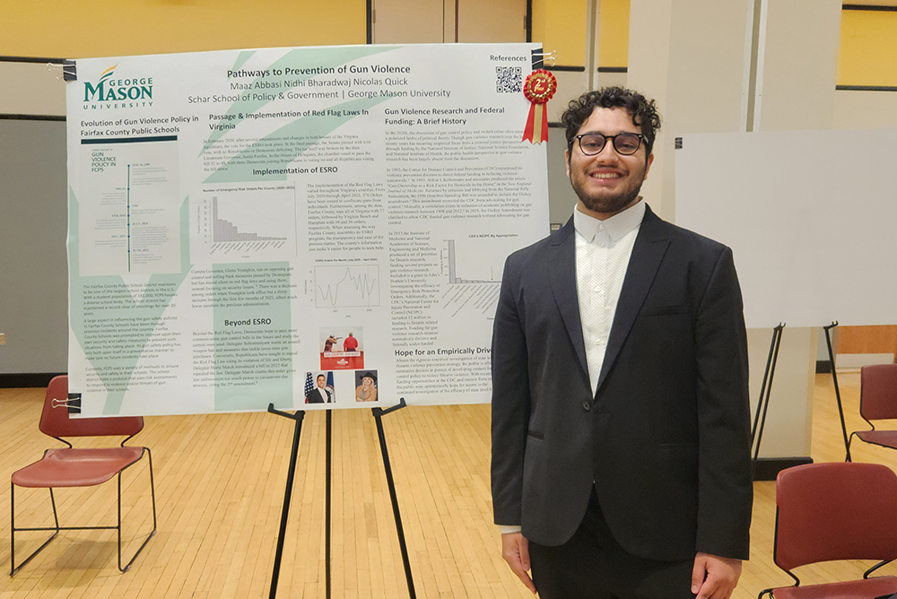 An IR Policy Task Force student with glasses in a black suit and white shirt standing in front of a poster on the Pathways to Prevention of Gun Violence.