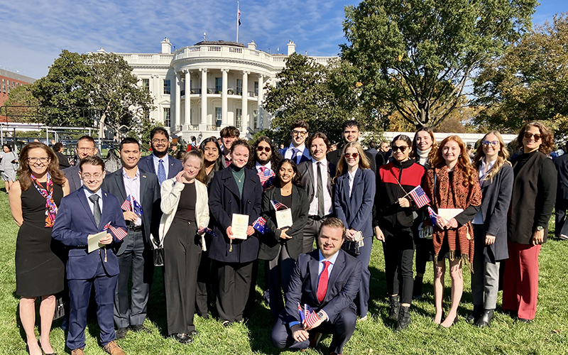 A large group of young people dress professionally stand in front of the White House in Washington, D.C.