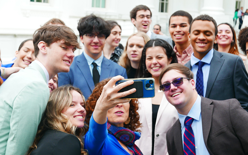 A woman in the foreground uses her cell phone to take a photo of herself and a group of young people in front of a white house.