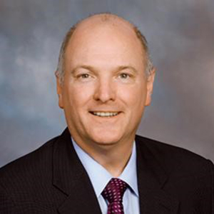 Terry Clower stands smiling while wearing a black suit, light blue shirt, and tie.