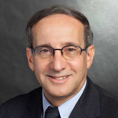 William J. Froehlich wearing a black suit, blue shirt, and a tie, sits and smiles.