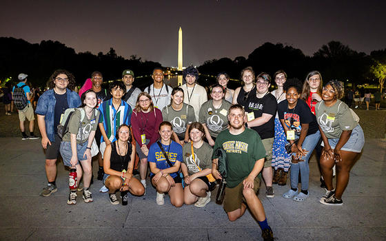 A group of teenagers in tee shirts stand pose with the lighted Washington Monument behind them.