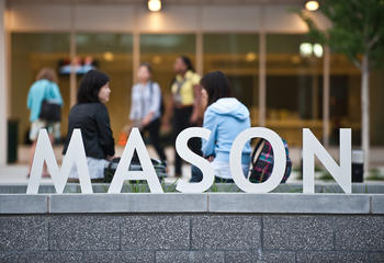 Silver letters spelling Mason sit on a wall outside of Van Metre Hall on Mason Square with students sitting and standing in the background.