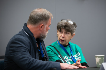 Louise Shelley wearing a teal t-shirt and with eyeglasses atop her head speaks to Patrick Kilbride who is wearing a gray jacket.